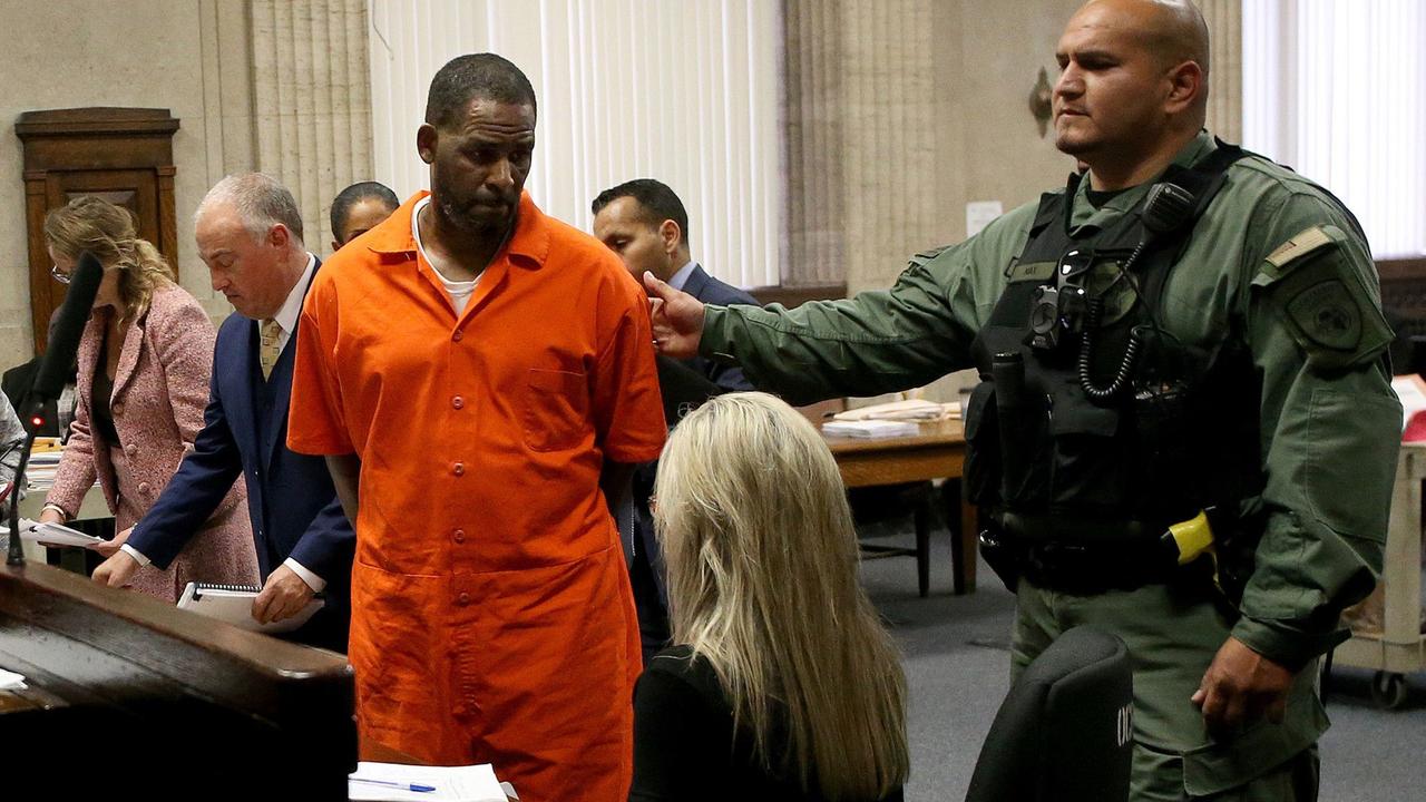 R. Kelly in einem orangen Ganzkörperanzug vor Gericht im Februar 2021. Ein Sicherheitsbeamter in Kampfanzug und schusssicherer Weste streckt seine Hand nach ihm aus.