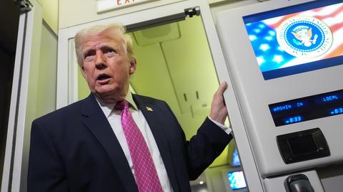 Das Bild zeigt Donald Trump an Bord seiner Präsidentenmaschine Air Force One. Er steht in einer Tür und spricht zu Journalisten.