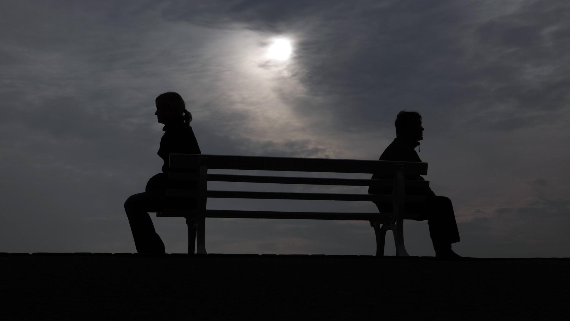Silhouette von einem Mann und einer Frau, die mit Abstand zu einander auf einer Ban sitzen