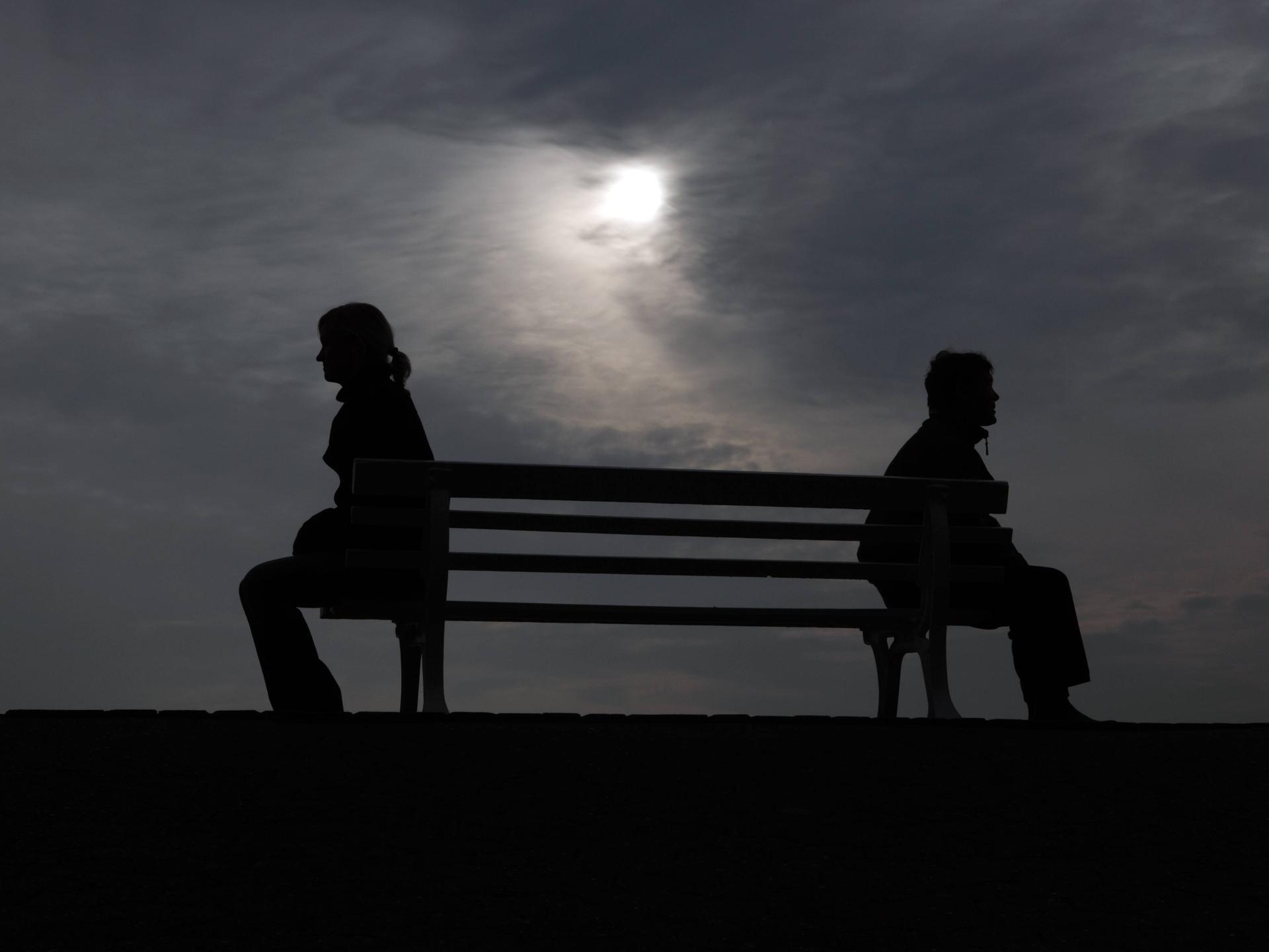 Silhouette von einem Mann und einer Frau, die mit Abstand zu einander auf einer Ban sitzen