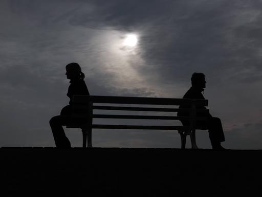 Silhouette von einem Mann und einer Frau, die mit Abstand zu einander auf einer Ban sitzen