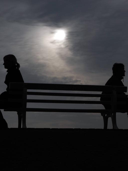 Silhouette von einem Mann und einer Frau, die mit Abstand zu einander auf einer Ban sitzen