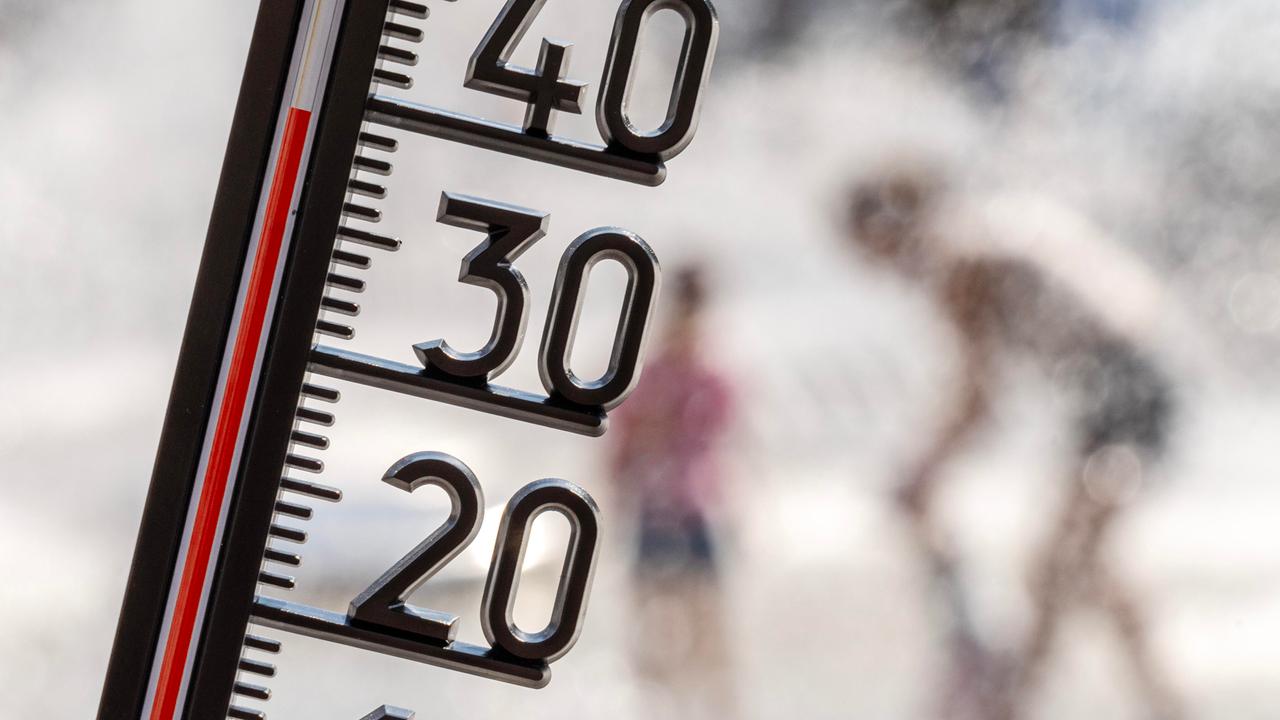 Ein Thermometer zeigt eine Temperatur von kanpp 40 Grad. Im Hintergrund sieht man unscharf Menschen, die sich unter einer Wasserfontäne abkühlen.