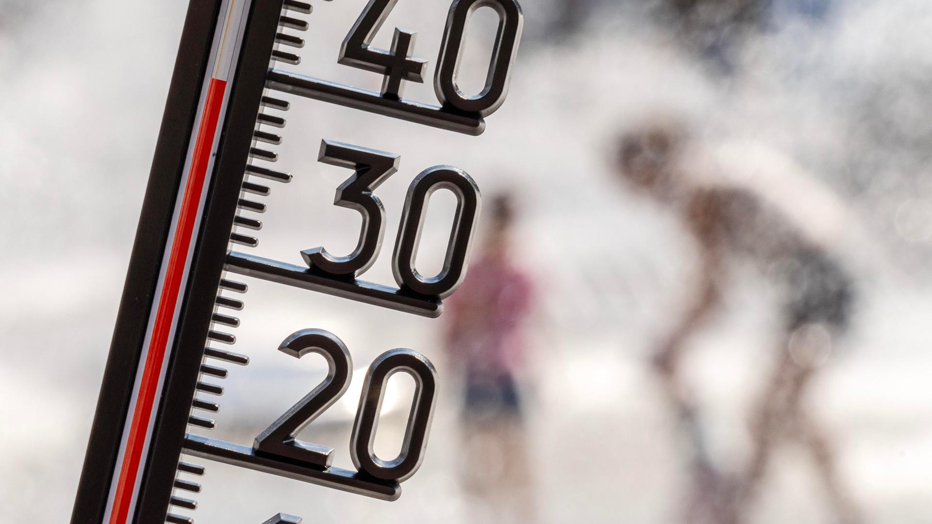 Ein Thermometer zeigt eine Temperatur von kanpp 40 Grad. Im Hintergrund sieht man unscharf Menschen, die sich unter einer Wasserfontäne abkühlen.