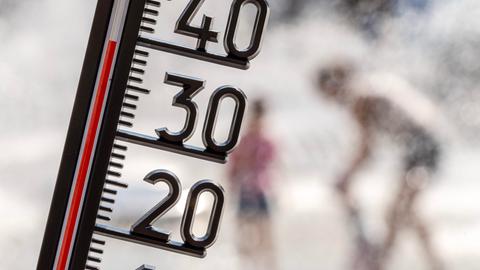 Ein Thermometer zeigt eine Temperatur von kanpp 40 Grad. Im Hintergrund sieht man unscharf Menschen, die sich unter einer Wasserfontäne abkühlen.