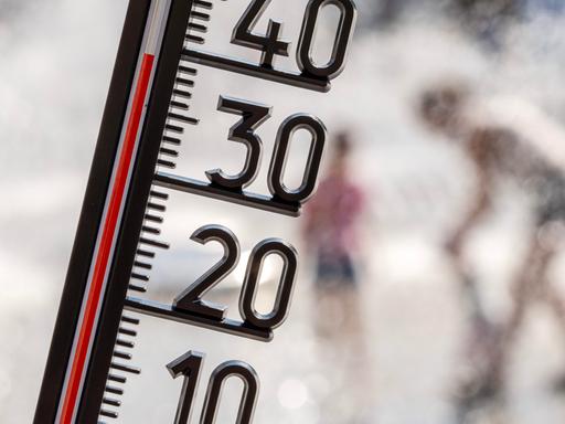 Ein Thermometer zeigt eine Temperatur von kanpp 40 Grad. Im Hintergrund sieht man unscharf Menschen, die sich unter einer Wasserfontäne abkühlen.
