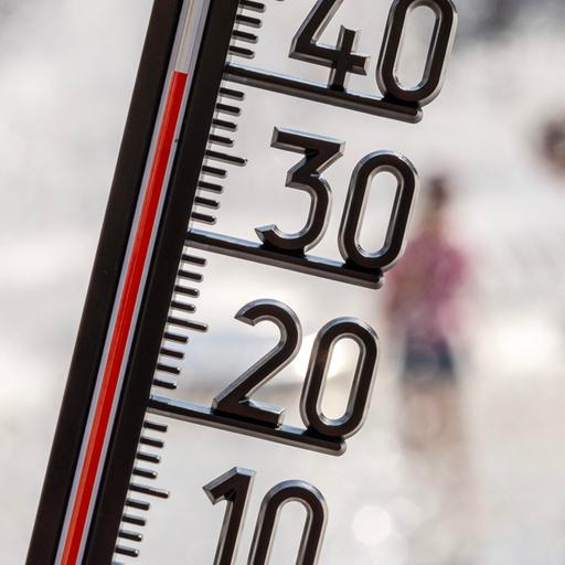 Ein Thermometer zeigt eine Temperatur von kapp 40 Grad, im Hintergrund sieht man in der Unschärfe Meschen, die sich unter einer Wasserfontäne abkühlen.