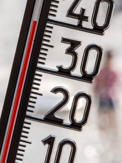 Ein Thermometer zeigt eine Temperatur von kanpp 40 Grad. Im Hintergrund sieht man unscharf Menschen, die sich unter einer Wasserfontäne abkühlen.