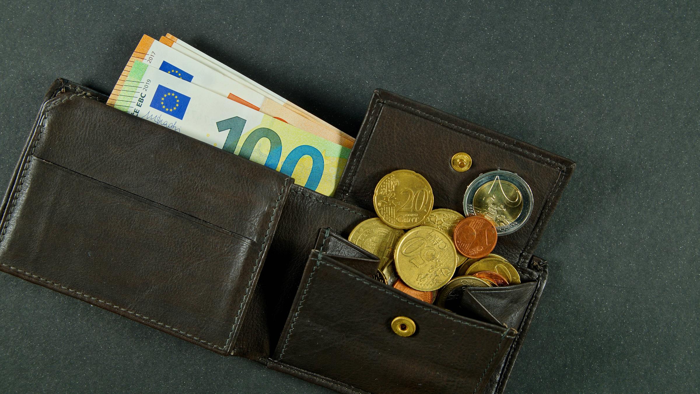 Eine dunkle, geöffnete Ledergeldbörse. Einige Euroscheine sind herausgezogen, Münzen sind aus dem geöffneten Münzfach gerutscht.