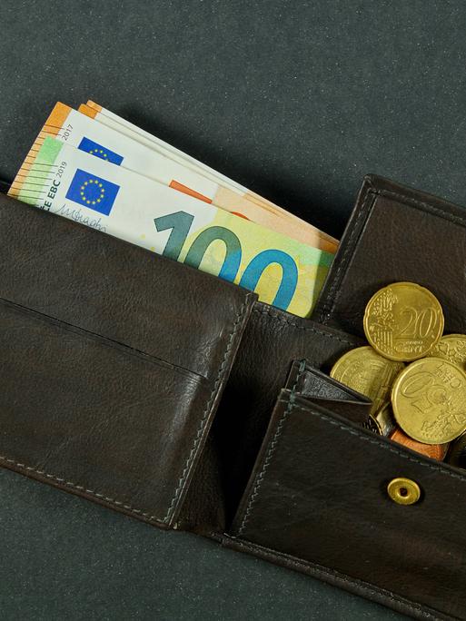 Eine dunkle, geöffnete Ledergeldbörse. Einige Euroscheine sind herausgezogen, Münzen sind aus dem geöffneten Münzfach gerutscht.