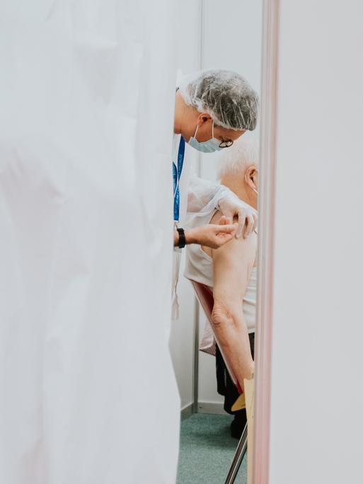 Ein älterer, weißhaariger Mann wird, hinter einem Vorhang, von einem Arzt mit Mundschutz in die Schulter geimpft.