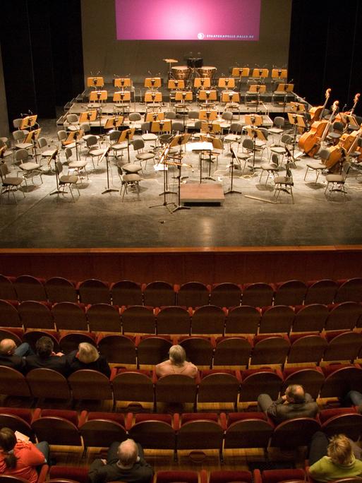 Publikum sitzt vor einer Bühne mit leeren Orchesterstühlen.
