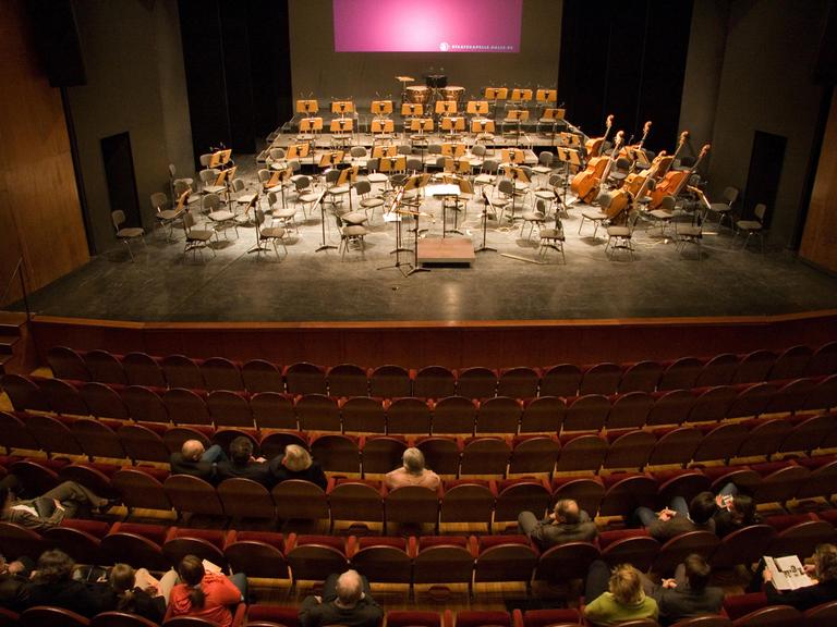 Publikum sitzt vor einer Bühne mit leeren Orchesterstühlen.