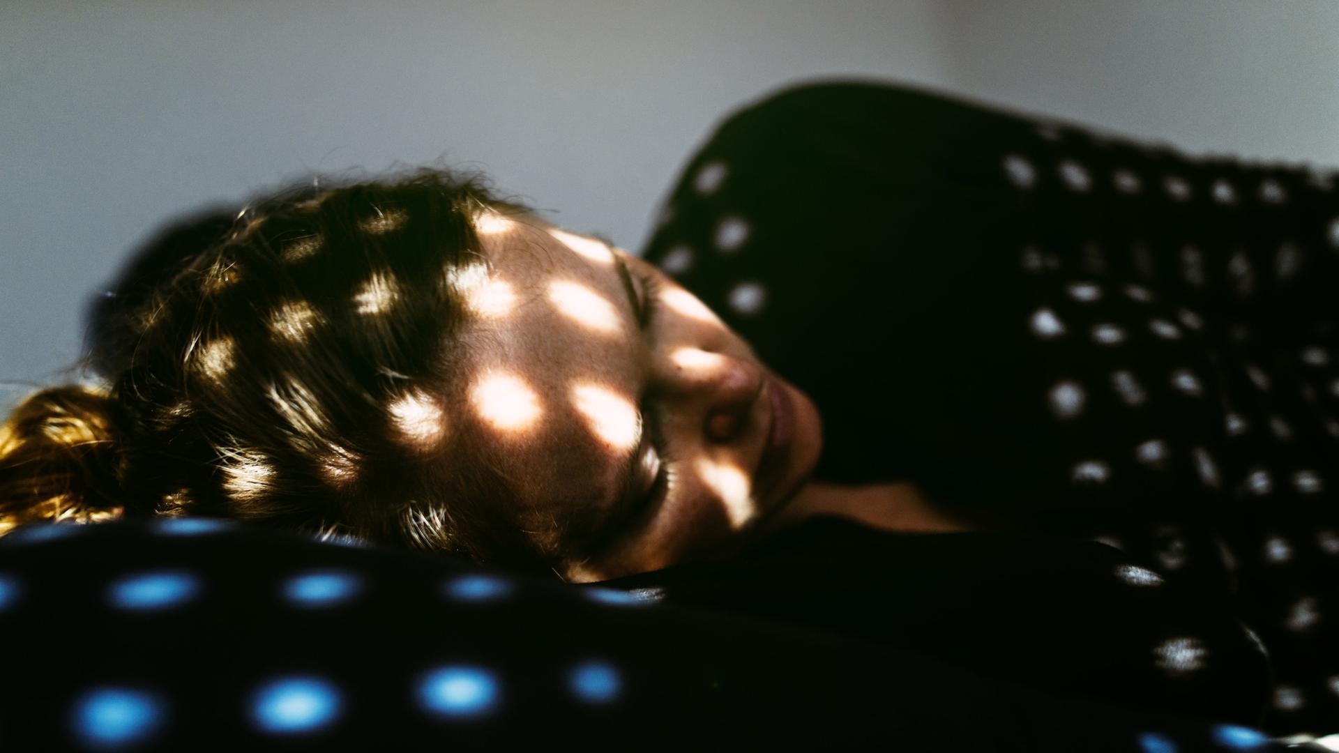 Eine Frau liegt in Fötusstellung auf einem Bett, Sonnensprenkel fallen durch eine halb geschlossene Jalousie auf sie.