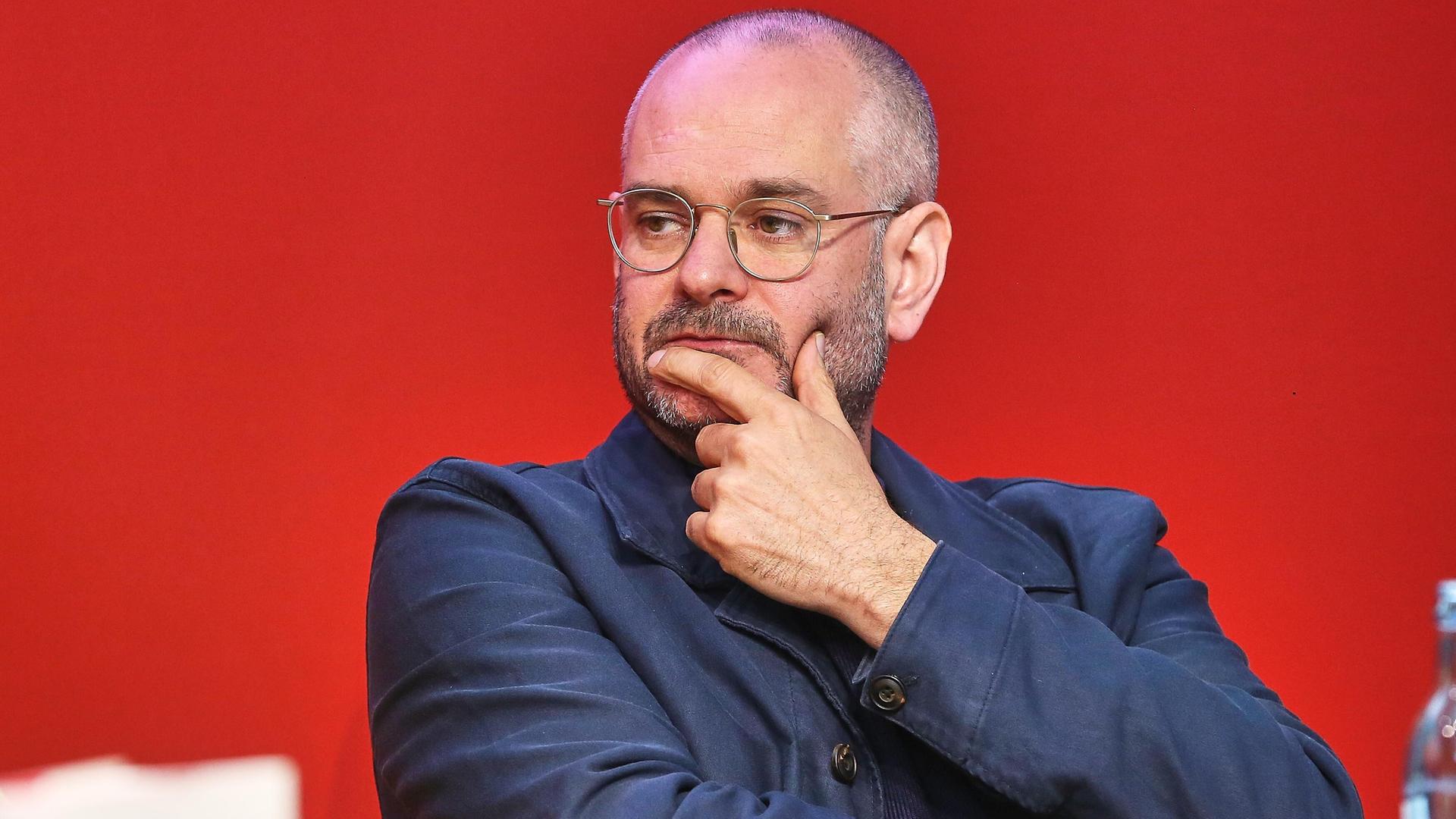 Der Schriftsteller Jonas Lüscher sitzt auf einem Podium und hält sich mit der linken Hand das Kinn. 