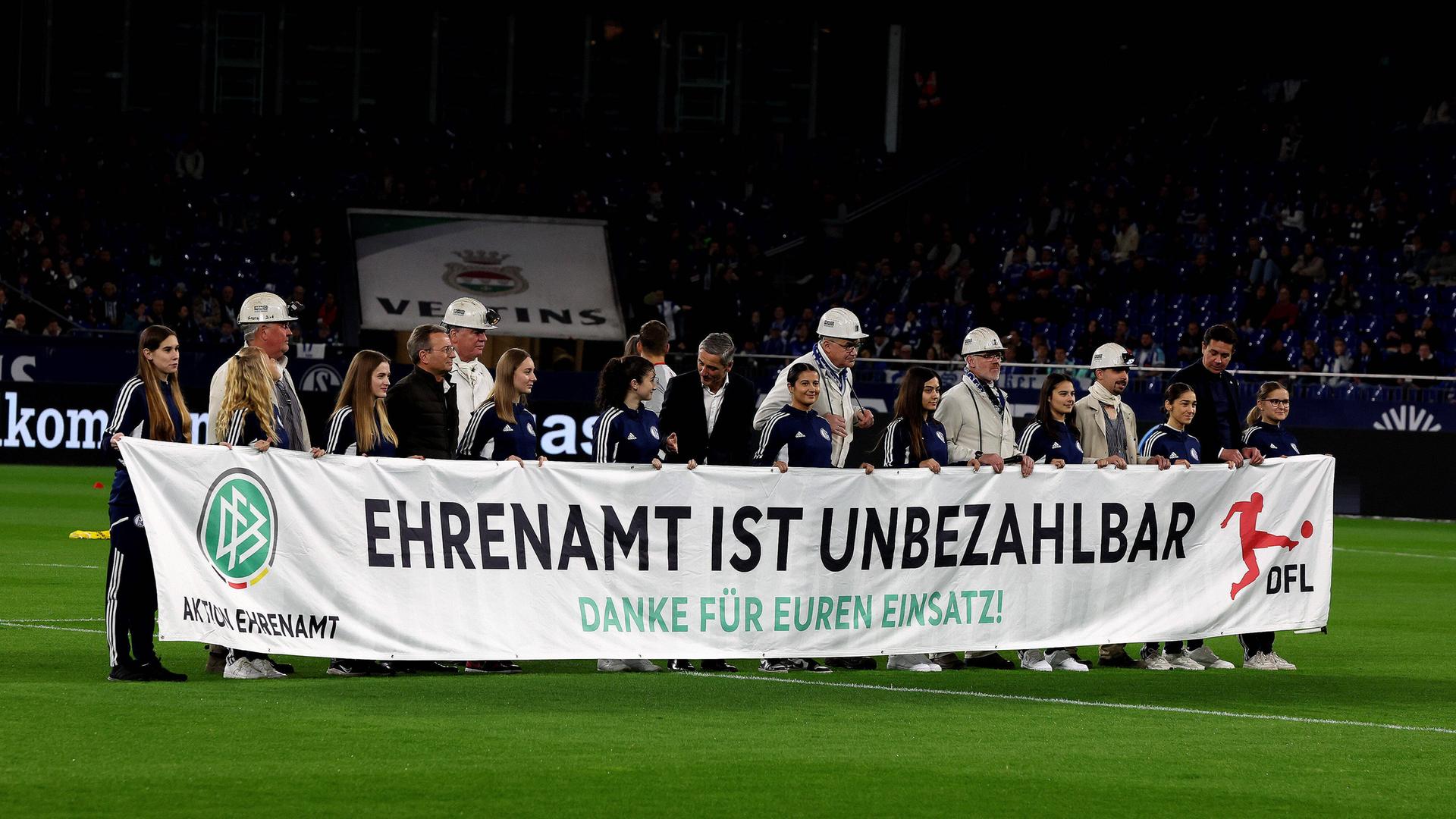 Menschen halten ein Banner mit der Aufschrift "Ehrenamt ist unbezahlbar" vor dem Bundesliga-Spiel zwischen dem FC Schalke 04 und dem 1. FSV Mainz 05.