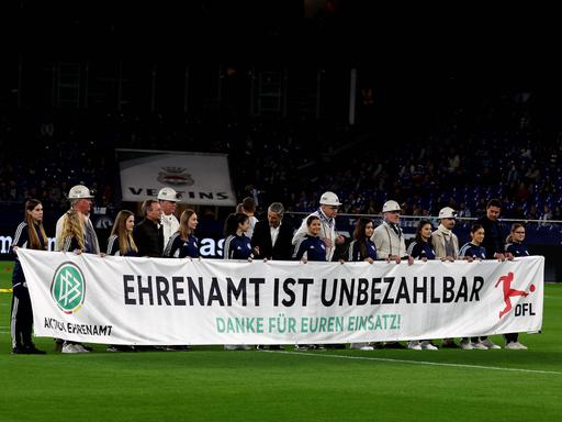 Menschen halten ein Banner mit der Aufschrift "Ehrenamt ist unbezahlbar" vor dem Bundesliga-Spiel zwischen dem FC Schalke 04 und dem 1. FSV Mainz 05.