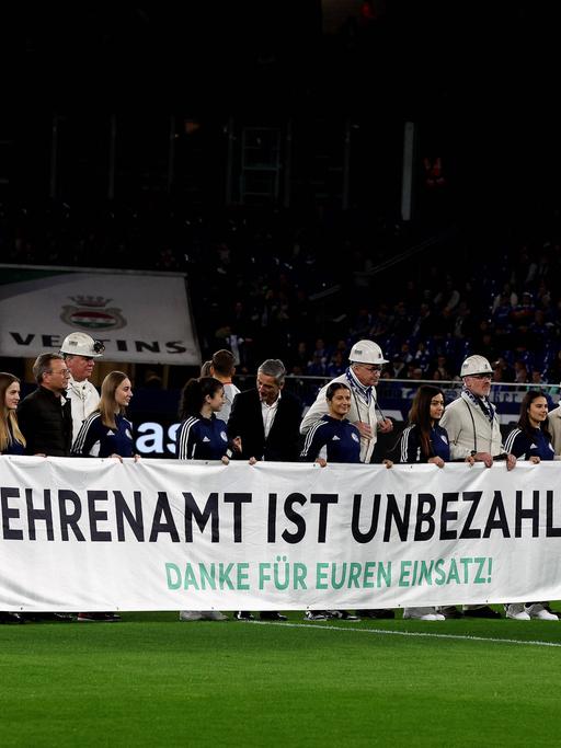 Menschen halten ein Banner mit der Aufschrift "Ehrenamt ist unbezahlbar" vor dem Bundesliga-Spiel zwischen dem FC Schalke 04 und dem 1. FSV Mainz 05.
