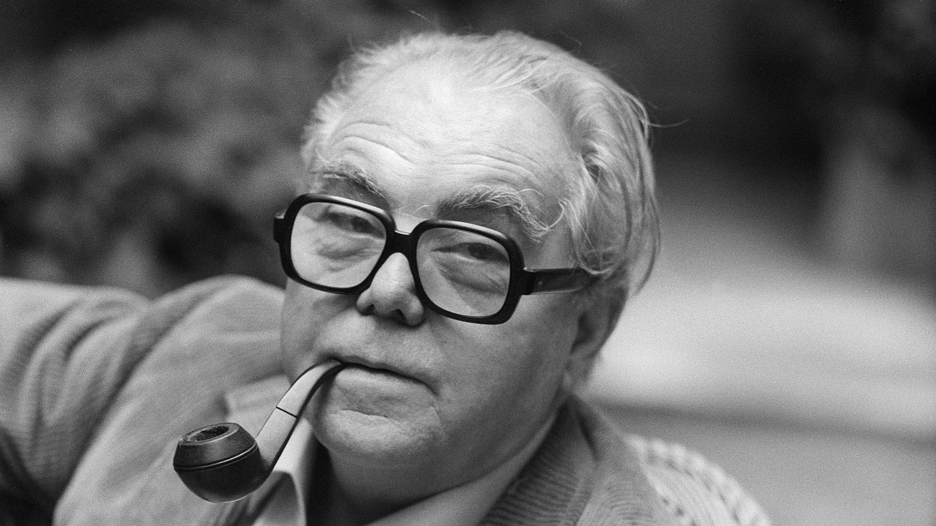 Porträt von Max Frisch in schwarz-weiß. Er hat eine Tabakpfeiffe im Mund und trägt eine Brille mit dickem, dunklen Rand. Er hat ein helles Hemd an und trägt eine helle Cordjacke.