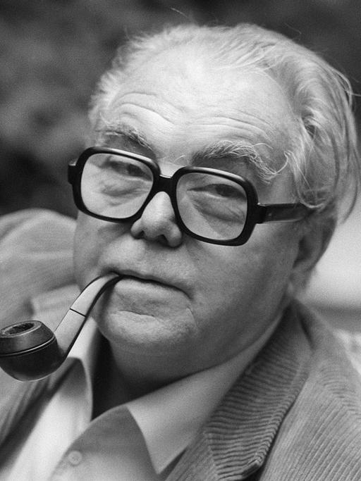Porträt von Max Frisch in schwarz-weiß. Er hat eine Tabakpfeiffe im Mund und trägt eine Brille mit dickem, dunklen Rand. Er hat ein helles Hemd an und trägt eine helle Cordjacke.