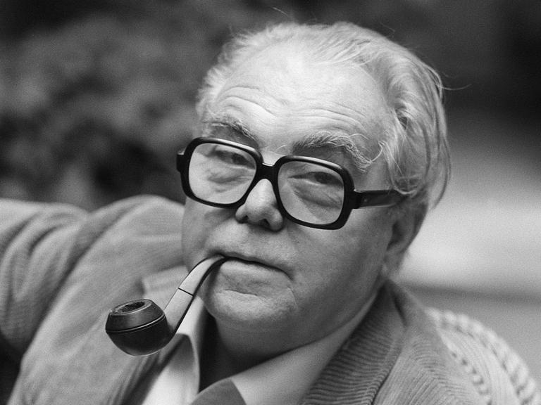 Porträt von Max Frisch in schwarz-weiß. Er hat eine Tabakpfeiffe im Mund und trägt eine Brille mit dickem, dunklen Rand. Er hat ein helles Hemd an und trägt eine helle Cordjacke.