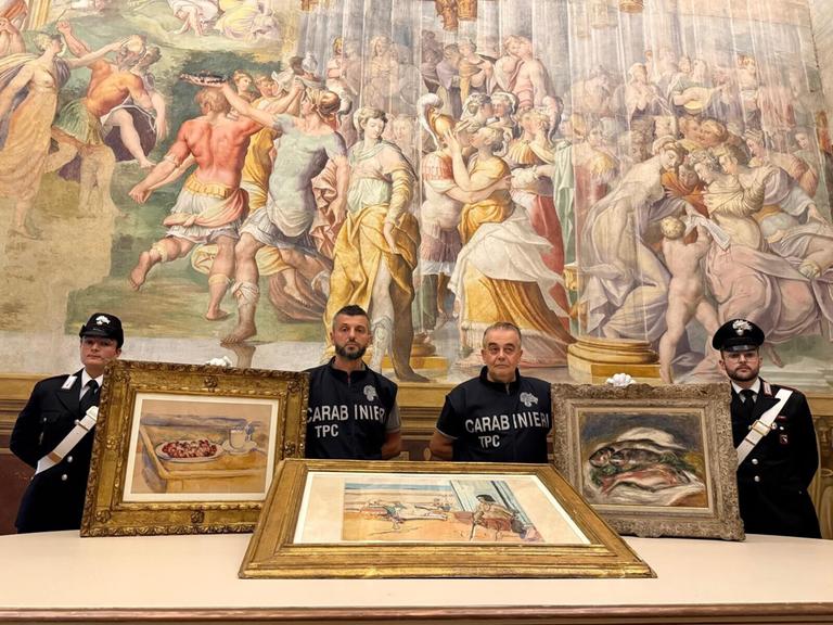Vier Polizisten stehen vor einem farbigen Wandteppich, vor ihnen liegen auf einem Tisch drei Gemälde mit breiten Rahmen versehen