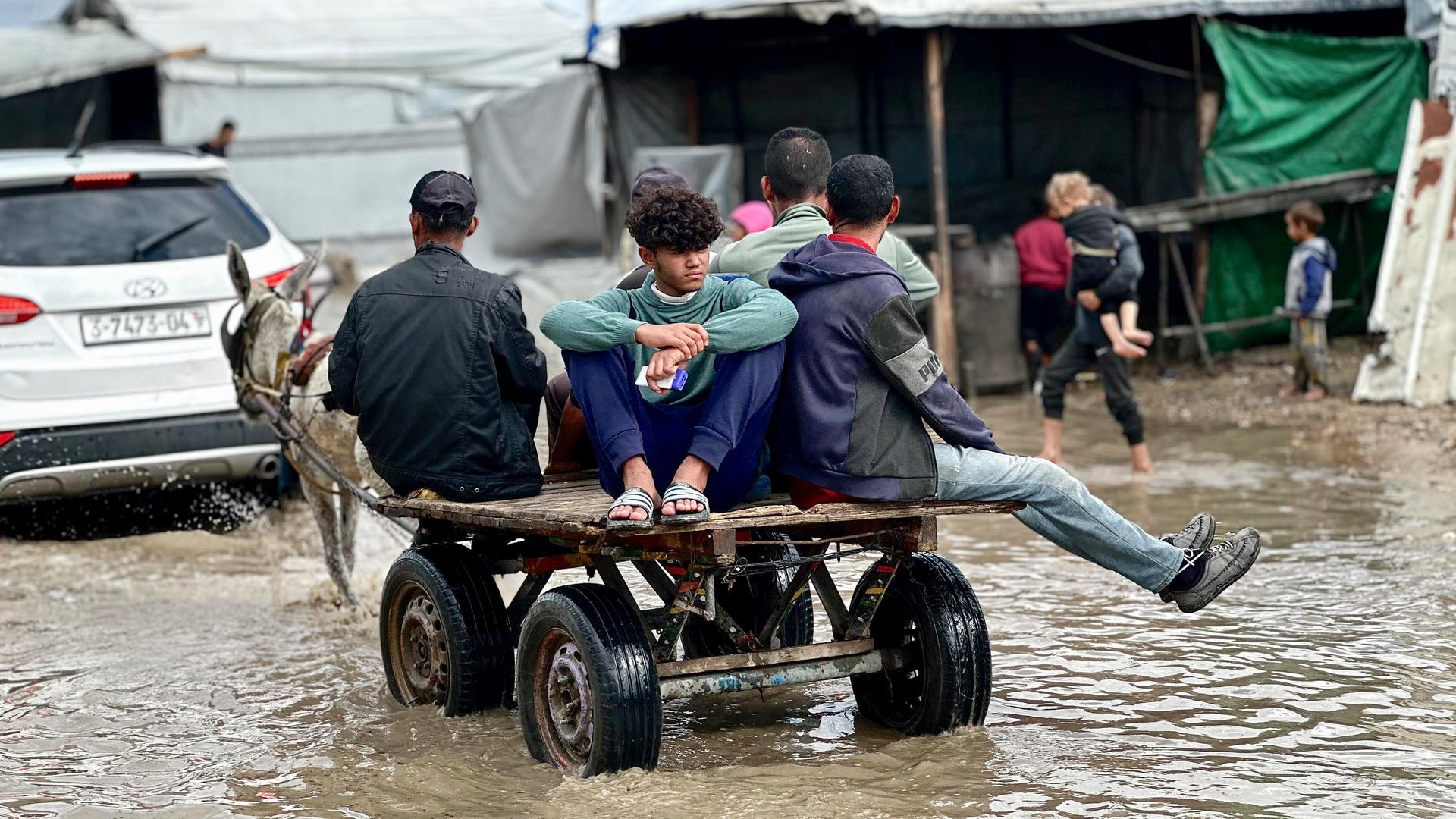 Eine Gruppe junger Männer sitzt auf einem kleinen Gefährt und fährt durch wadenhohes schlammiges Wasser. Im Hintergrund läuft eine Mutter mit kleinen Kindern.