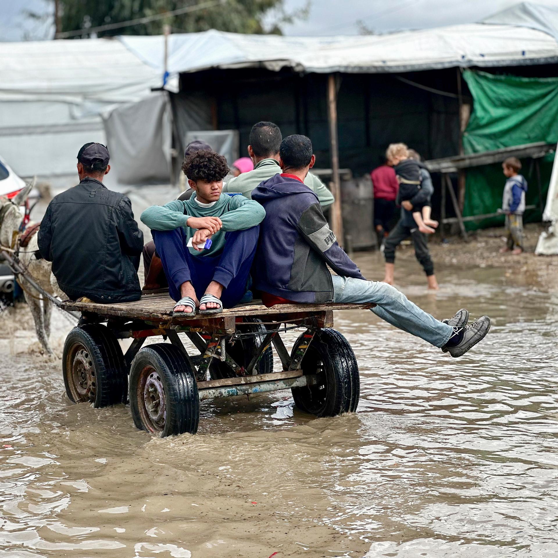 Eine Gruppe junger Männer sitzt auf einem kleinen Gefährt und fährt durch wadenhohes schlammiges Wasser. Im Hintergrund läuft eine Mutter mit kleinen Kindern.