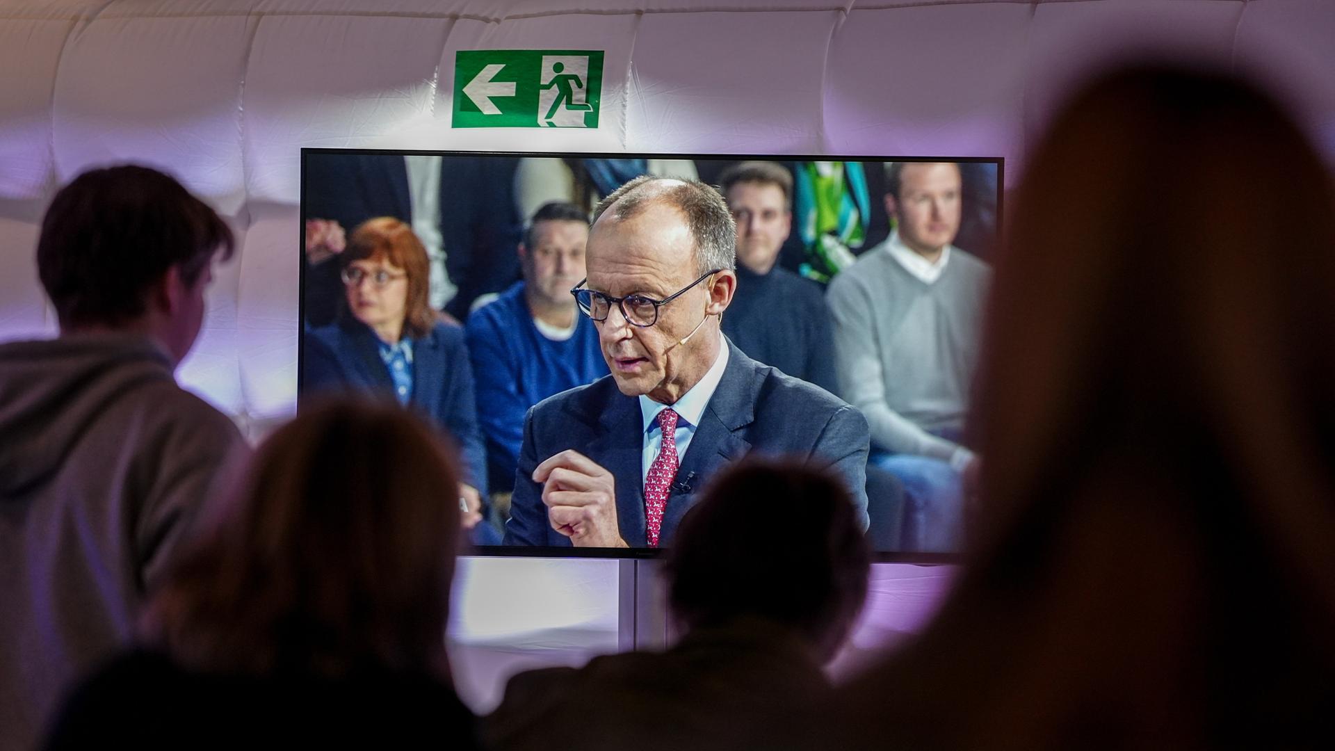 Friedrich Merz ist auf einem Fernseher zu sehen. Vor dem Fernseher, im Anschnitt, sind unscharf zuschauende Personen zu sehen.