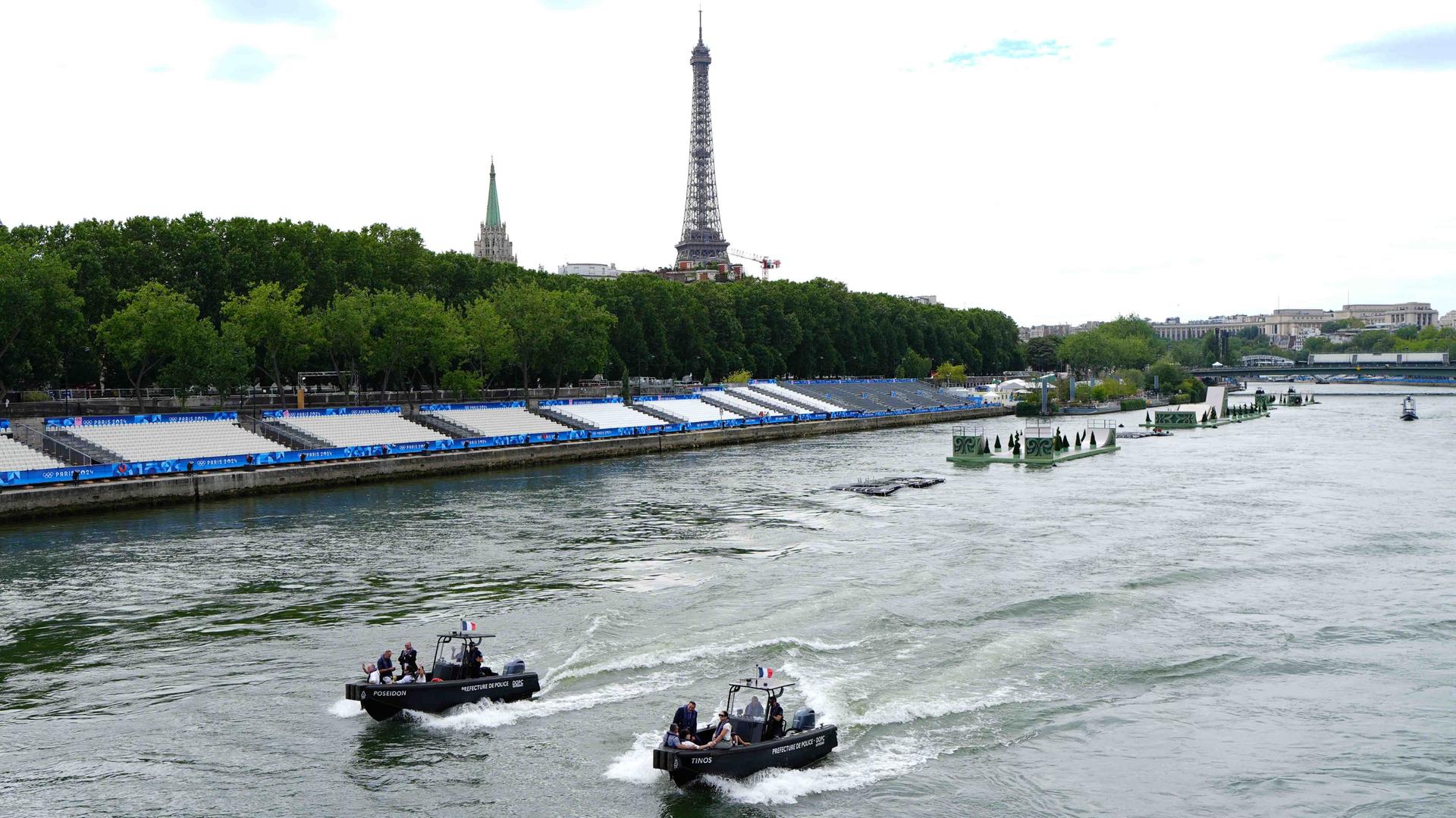 Man sieht den Fluss Seine. Darauf fahren zwei Polizeiboote. Am Ufer stehen zahlreiche leere Tribünen. Im Hintergrund ist der Eifelturm zu sehen.