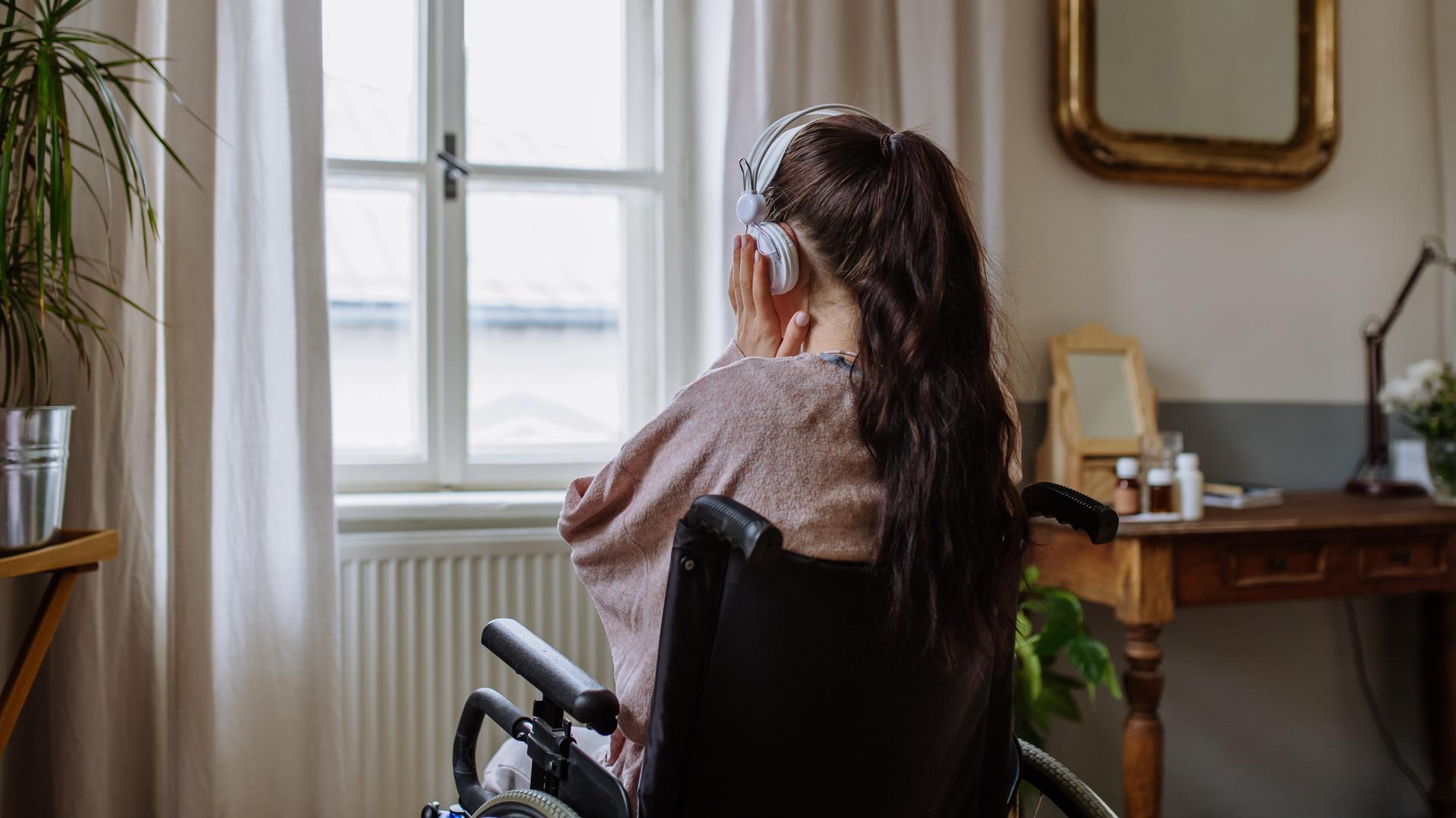 Ein Teenager-Mädchen sitzt in einem Rollstuhl, hat den Rücken der Kamera zugewandt und hört Musik, während sie aus dem Fenster schaut.