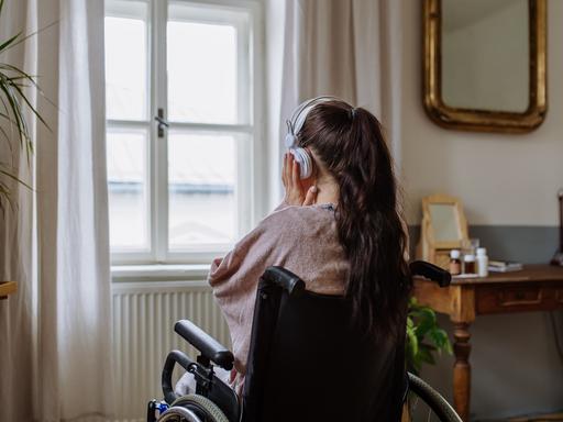 Ein Teenager-Mädchen sitzt in einem Rollstuhl, hat den Rücken der Kamera zugewandt und hört Musik, während sie aus dem Fenster schaut.