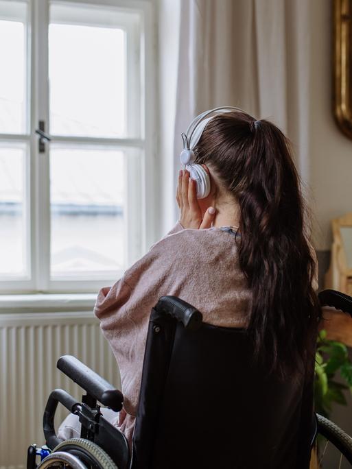 Ein Teenager-Mädchen sitzt in einem Rollstuhl, hat den Rücken der Kamera zugewandt und hört Musik, während sie aus dem Fenster schaut.