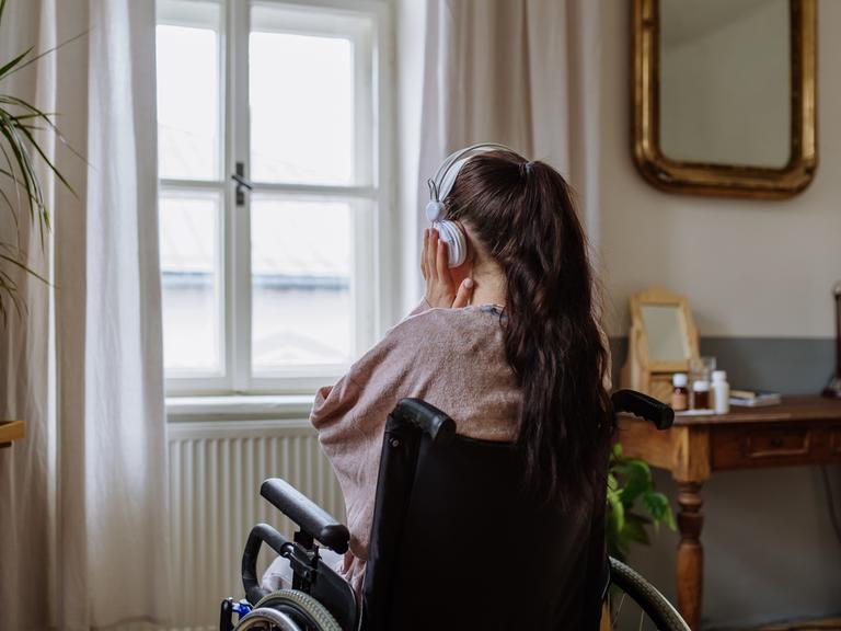 Ein Teenager-Mädchen sitzt in einem Rollstuhl, hat den Rücken der Kamera zugewandt und hört Musik, während sie aus dem Fenster schaut.