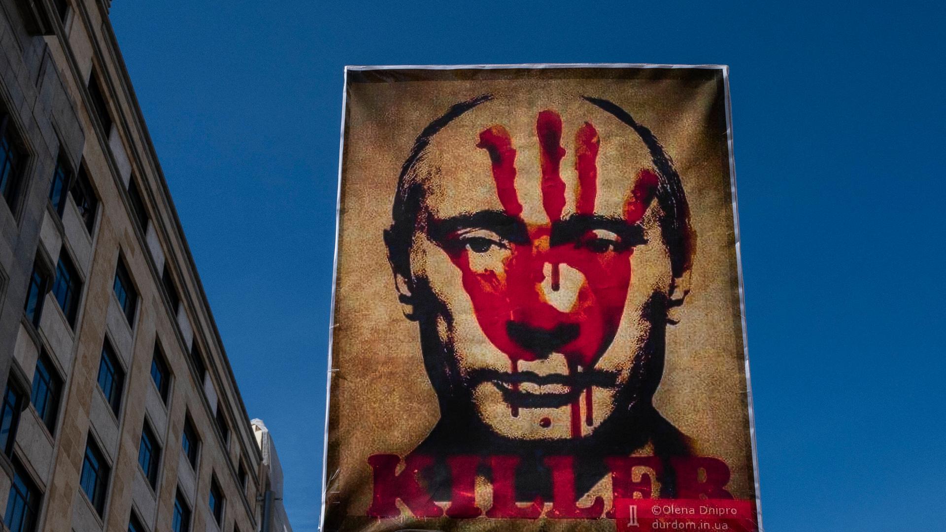 Ein Plakat mit Putins Porträt, das mit einem roten Handaufdruck und dem Schriftzug "Killer" versehen ist, wird durch eine Straße getragen.