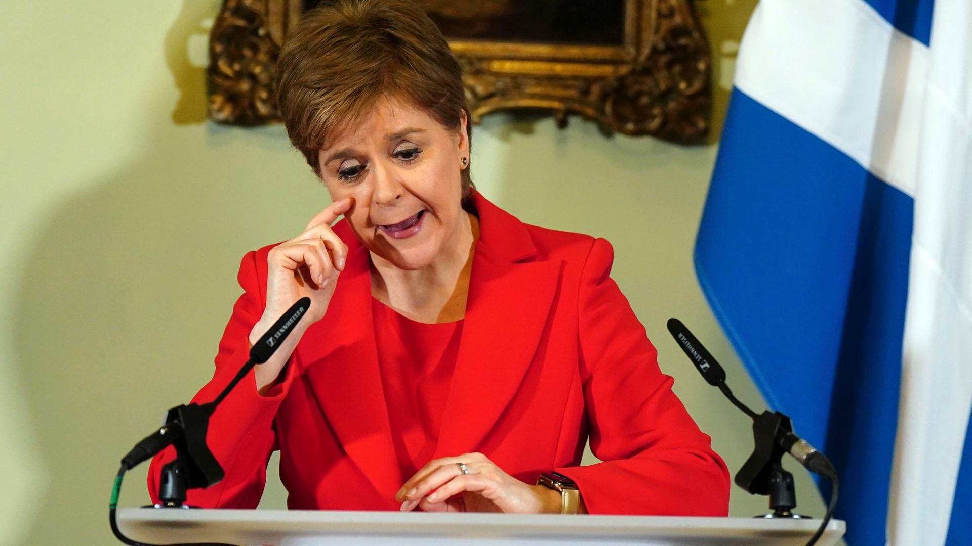 Nicola Sturgeon trägt einen roten Anzug. Sie steht hinter einem Pult und redet.