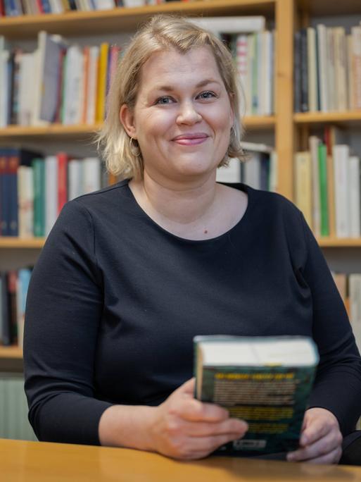 Die Schriftstellerin Berit Glanz zu Gast im Literaturhaus Schleswig-Holstein in Kiel. Sie lächelt in die Kamera, sitzt hinter einem Schreibtisch und hält ein Buch in der Hand. Im Hintergrund sind Bücherregale zu erkennen. Die Schriftstellerin Berit Glanz zu Gast im Literaturhaus Schleswig-Holstein in Kiel. Sie lächelt in die Kamera, sitzt hinter einem Schreibtisch und hält ein Buch in der Hand. Im Hintergrund sind Bücherregale zu erkennen.