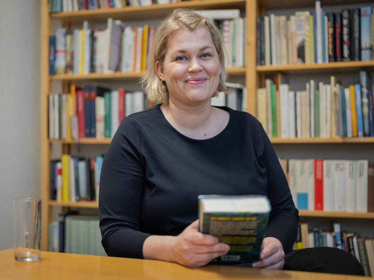 Die Schriftstellerin Berit Glanz zu Gast im Literaturhaus Schleswig-Holstein in Kiel. Sie lächelt in die Kamera, sitzt hinter einem Schreibtisch und hält ein Buch in der Hand. Im Hintergrund sind Bücherregale zu erkennen. Die Schriftstellerin Berit Glanz zu Gast im Literaturhaus Schleswig-Holstein in Kiel. Sie lächelt in die Kamera, sitzt hinter einem Schreibtisch und hält ein Buch in der Hand. Im Hintergrund sind Bücherregale zu erkennen.