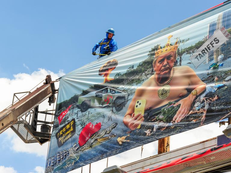 Arbeiter installieren eine Plakatwand der Künstlerin Karen Fiorito in Phoenix. Das Werk mit dem Titel „Swamp King“ kritisiert Donald Trumps Präsidentschaft mit Bildern, die Korruption, Autoritarismus und systemischen Verfall symbolisieren. 