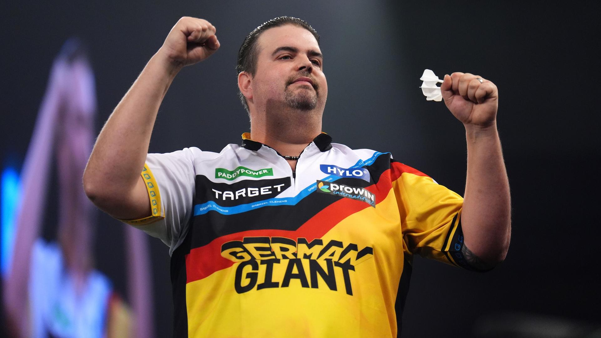 Gabriel Clemens trägt ein buntes Trikot. Er hält einen Dartpfeil in der Hand und hebt beide Hände zum Sieg.