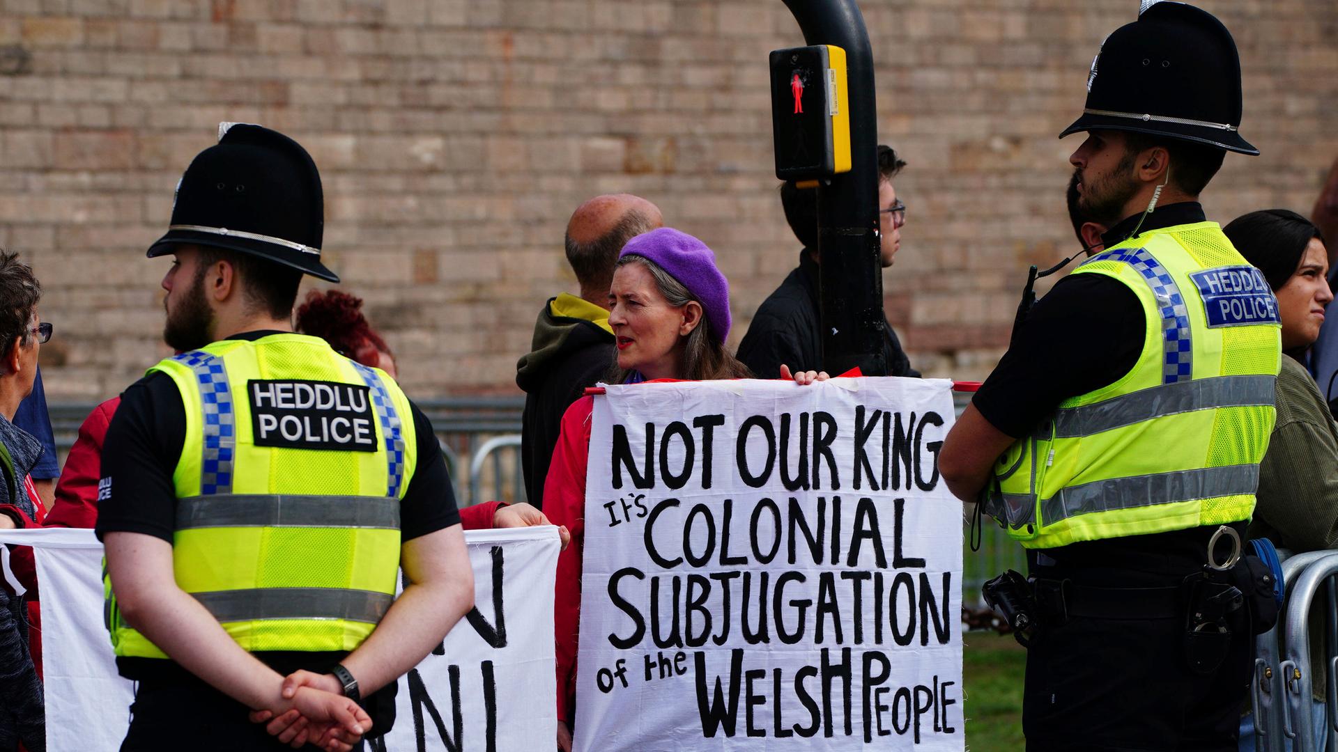 Eine Frau protestiert mit einem Schild gegen den neuen Königs George III. Darauf ist zu lesen: "Nicht unser König". Vor ihr stehen zwei britische Polizisten. 