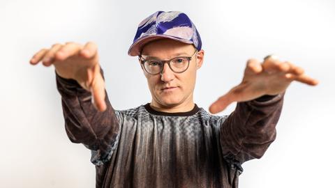 Der Musiker PeterLicht mit Kappe steht der Kamera gegenüber und streckt dabei Arme und Hände nach ihr aus.