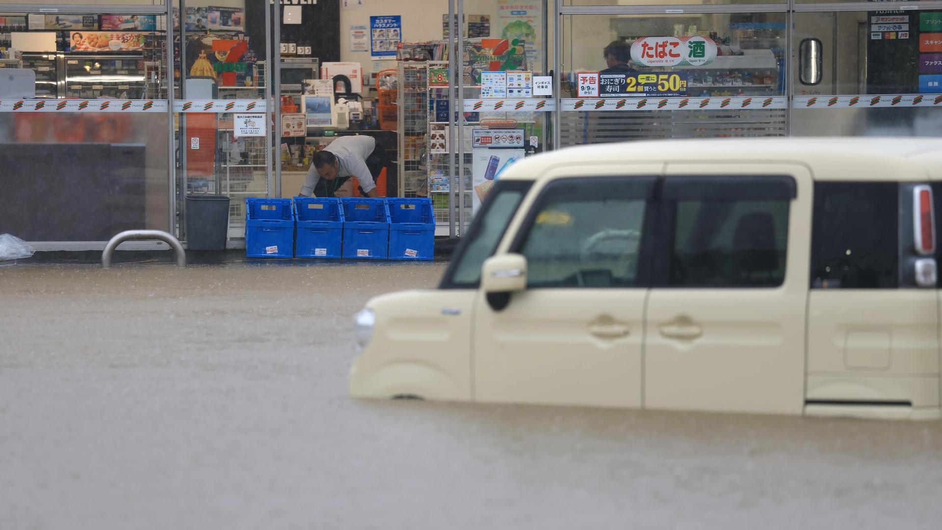 Ein Mann versucht, das Wasser aus seinem Laden zu schöpfen, im Vordergrund steht ein weißer Kleinbus, der bis über die Räder im Wasser steht. Ein Mann versucht, das Wasser aus seinem Laden zu schöpfen, im Vordergrund steht ein weißer Kleinbus, der bis über die Räder im Wasser steht.