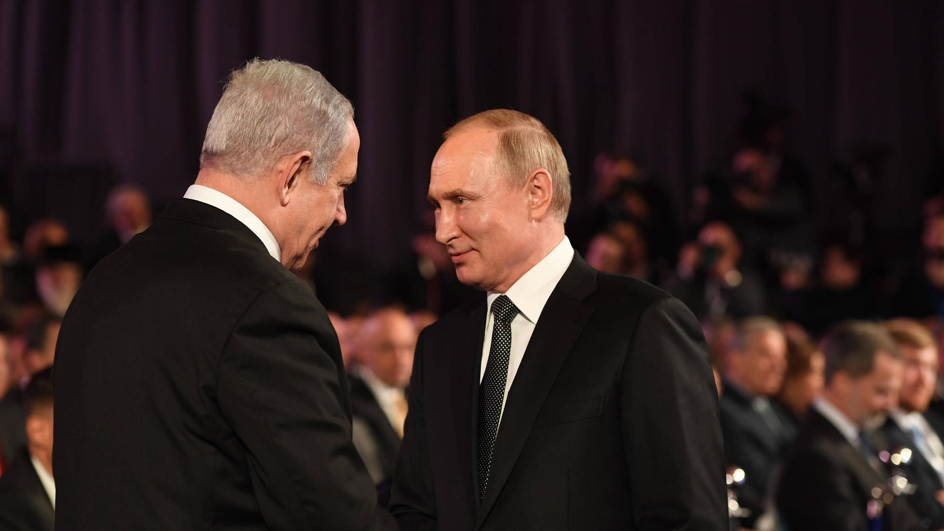 Benjamin Netanjahu und Wladimir Putin beim Händeschütteln vor schwarzem Hintergrund. Am unteren Rand des Bildes sind verschwommen Zuschauer zu erkennen.