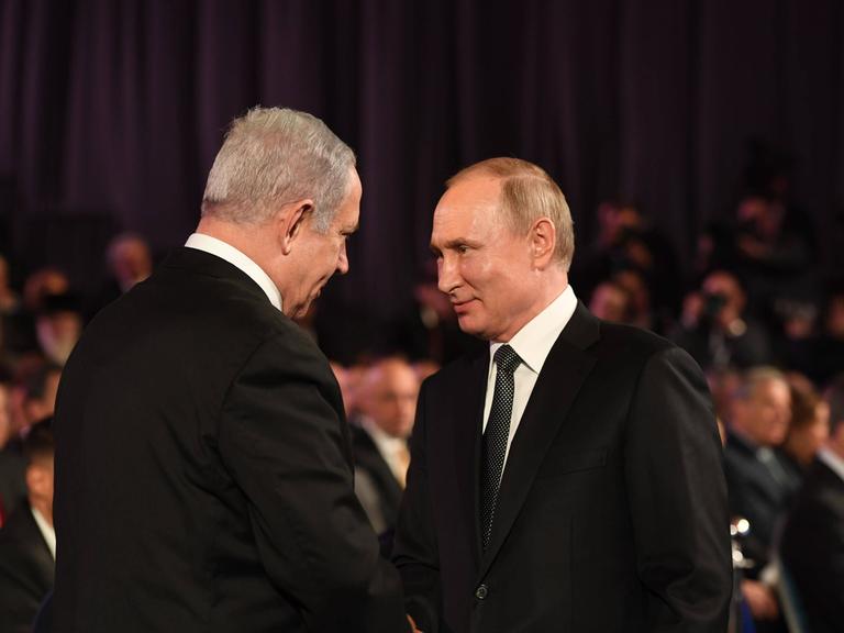 Benjamin Netanjahu und Wladimir Putin beim Händeschütteln vor schwarzem Hintergrund. Am unteren Rand des Bildes sind verschwommen Zuschauer zu erkennen.
