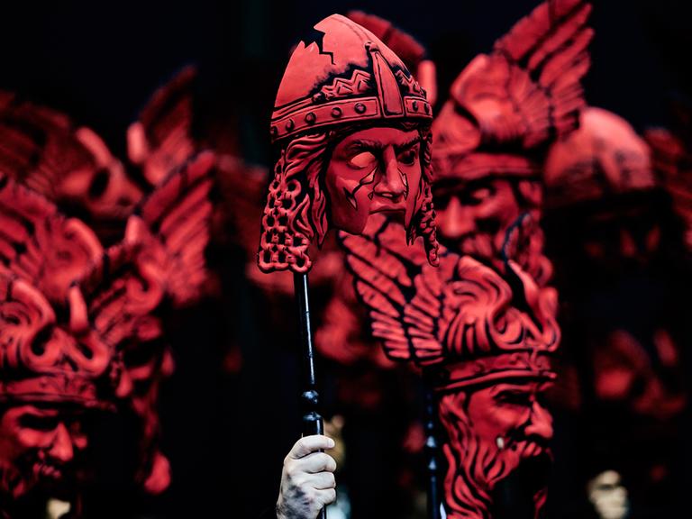 Auf der Bühne halten Darsteller bei der "Götterdämmerung" rote Masken in die Höhe.
