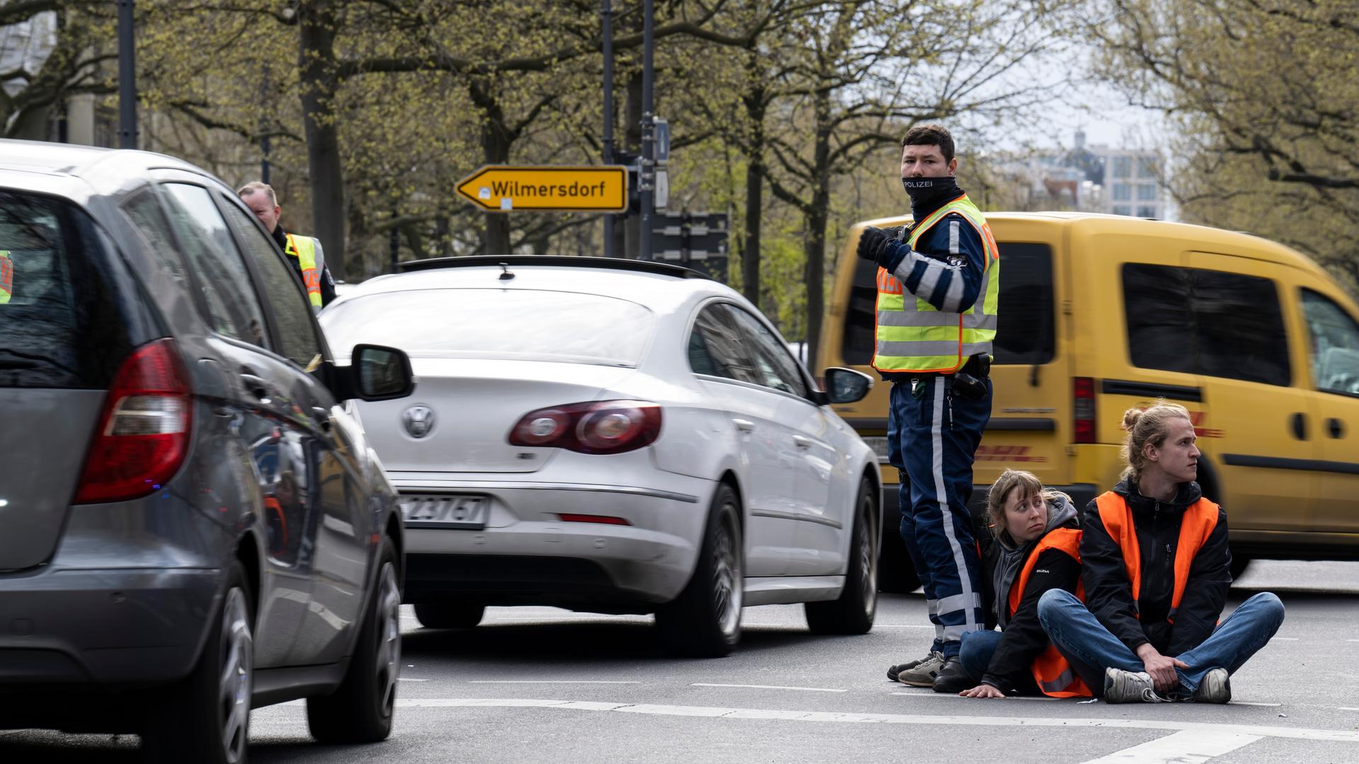 Zwei Aktivisten der Gruppe "Letzte Generation" sitzen auf dem Kurfürstendamm und werden von einem Polizisten vom Verkehr abgeschirmt.