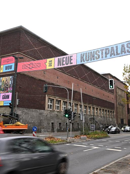 Quer über die Straße gespannte Banner weisen die Passanten 2023 auf die Wiedereröffnung des Kunstpalastes hin. Am Straßenrand sieht man das Gebäude des Kunstpalastes. 