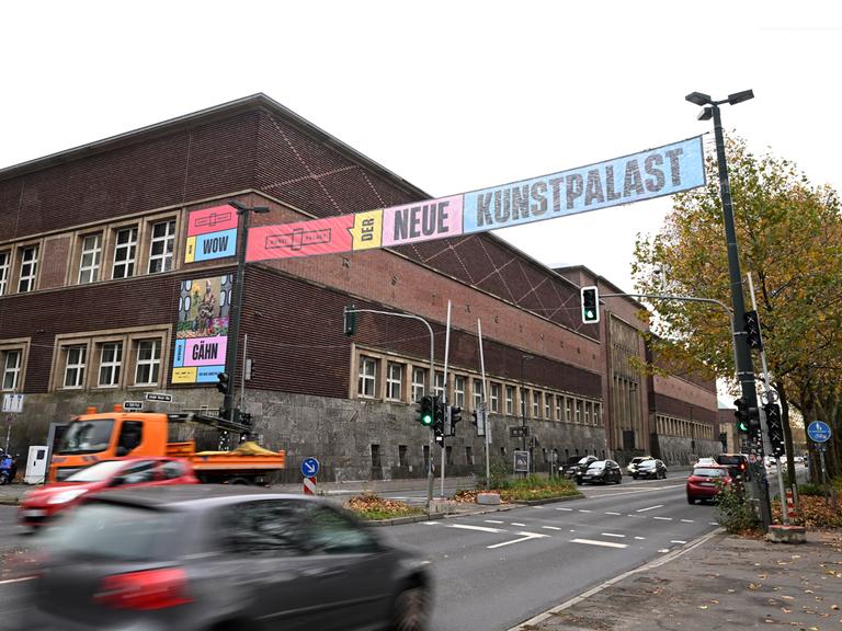 Quer über die Straße gespannte Banner weisen die Passanten 2023 auf die Wiedereröffnung des Kunstpalastes hin. Am Straßenrand sieht man das Gebäude des Kunstpalastes. 