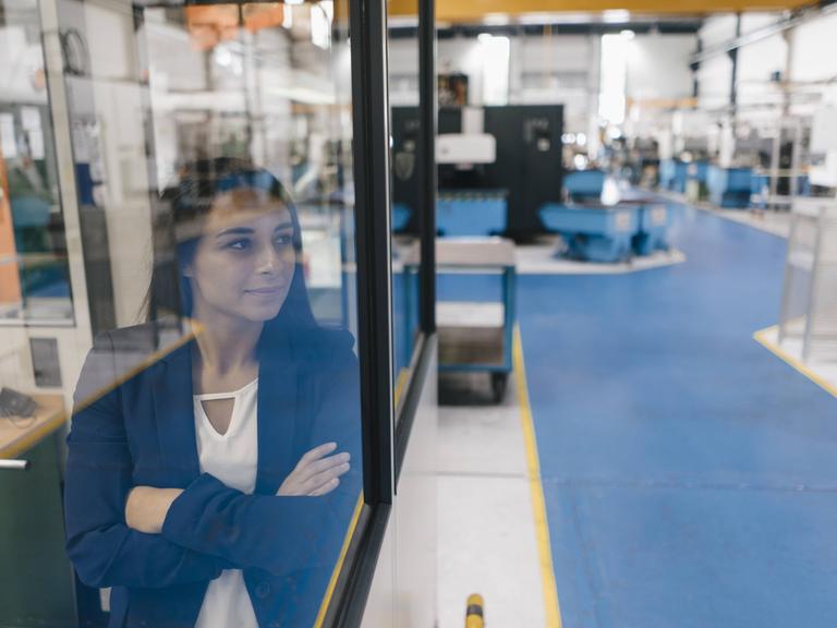 Eine junge Frau blickt zuversichtlich durch eine Scheibe in die Lagerhalle einer Firma. (Symbolfoto)